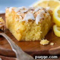A slice of almond flour lemon cake with a bite taken out, accompanied by fresh lemons on a white plate.