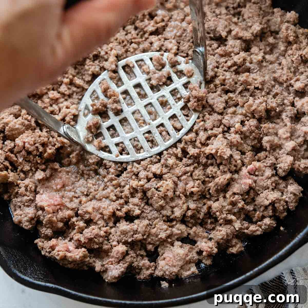 Revolutionary Gluten Free Weeknight Tacos 5 A wire masher actively breaking up ground beef in a skillet, ensuring a finely textured and uniform taco filling.