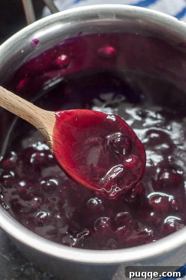 Homemade 10-Minute Maple Blueberry Syrup 4 A wooden spoon lifting blueberry syrup from a saucepan, showing its thickening texture.