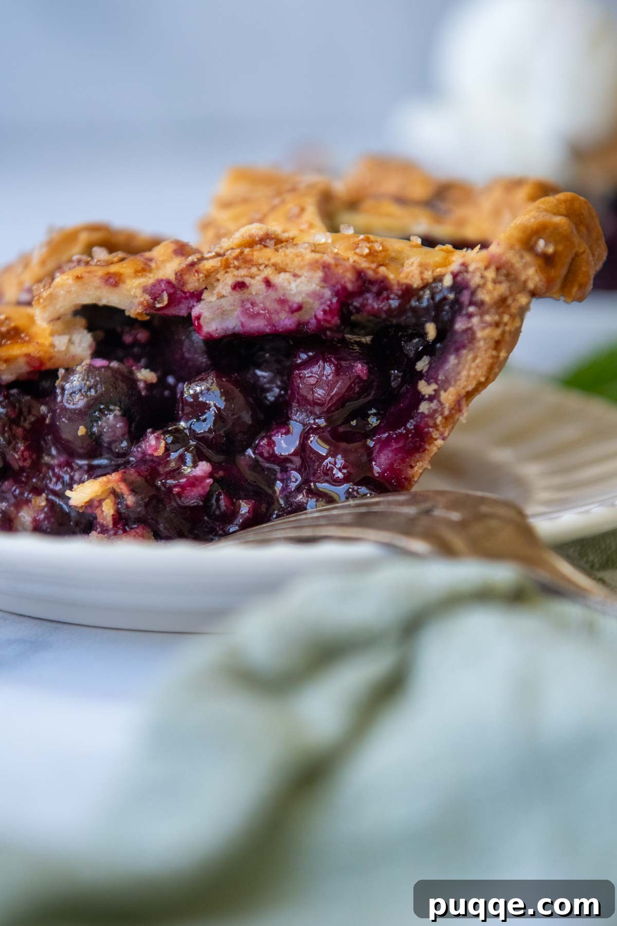 A close-up side view of a slice of gluten-free blueberry pie on a white plate, showcasing the flaky crust and juicy filling.
