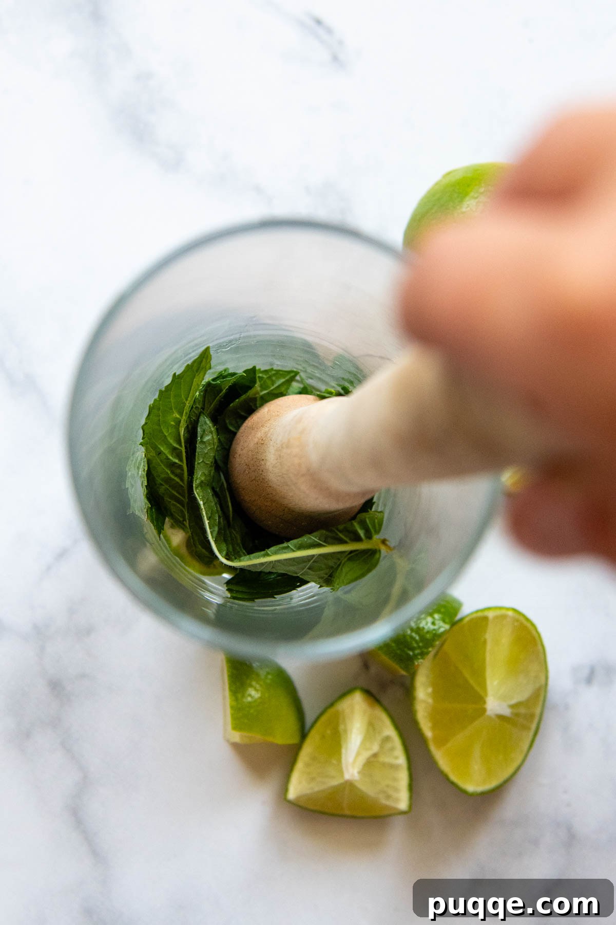 Close-up shot of a muddler gently pressing fresh mint and lime wedges at the bottom of a cocktail glass.