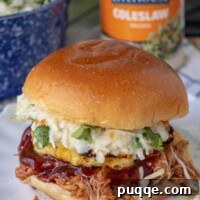 Quick Prep Slow Cooker BBQ Pulled Chicken 7 A BBQ pulled chicken sandwich on a plate with coleslaw, BBQ sauce, and grilled pineapple
