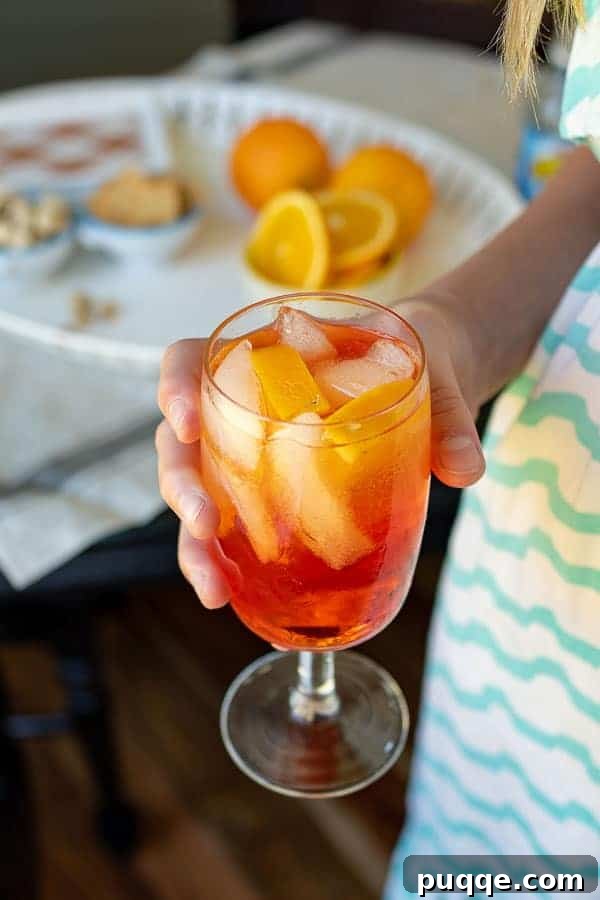 The Definitive Aperol Spritz Recipe 2 A hand holding a vibrant orange Aperol Spritz cocktail, glistening in the light, with a fresh orange slice garnish.