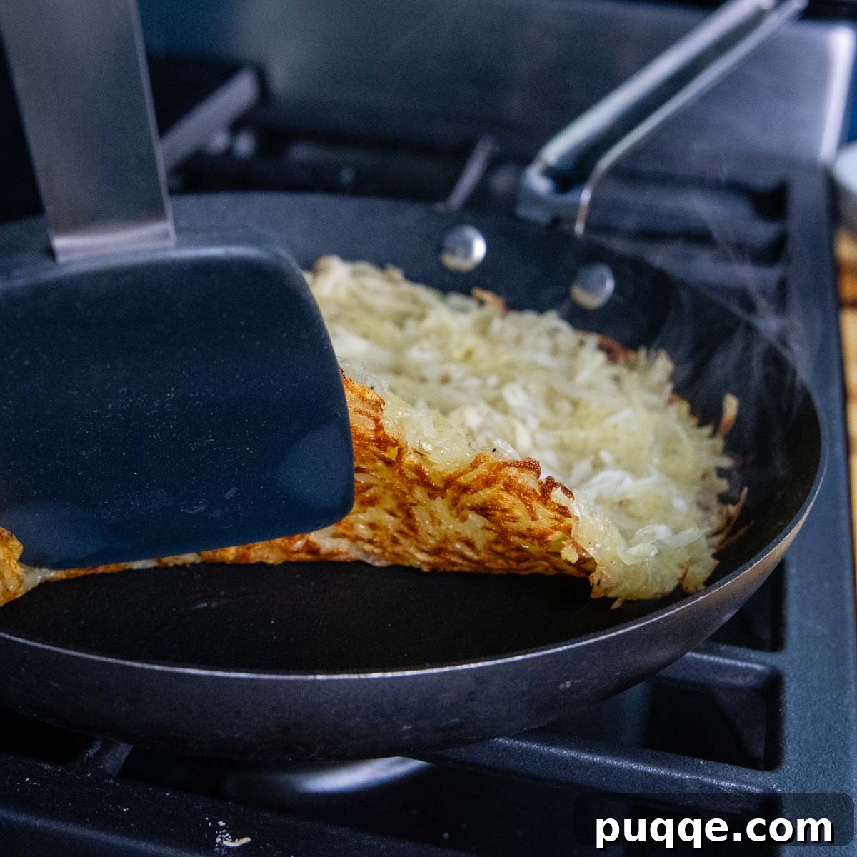 Effortless Crispy Potato Rösti 6 A spatula lifting the edge of a potato rosti in a skillet, revealing the beautifully browned and crispy underside, ready for flipping.