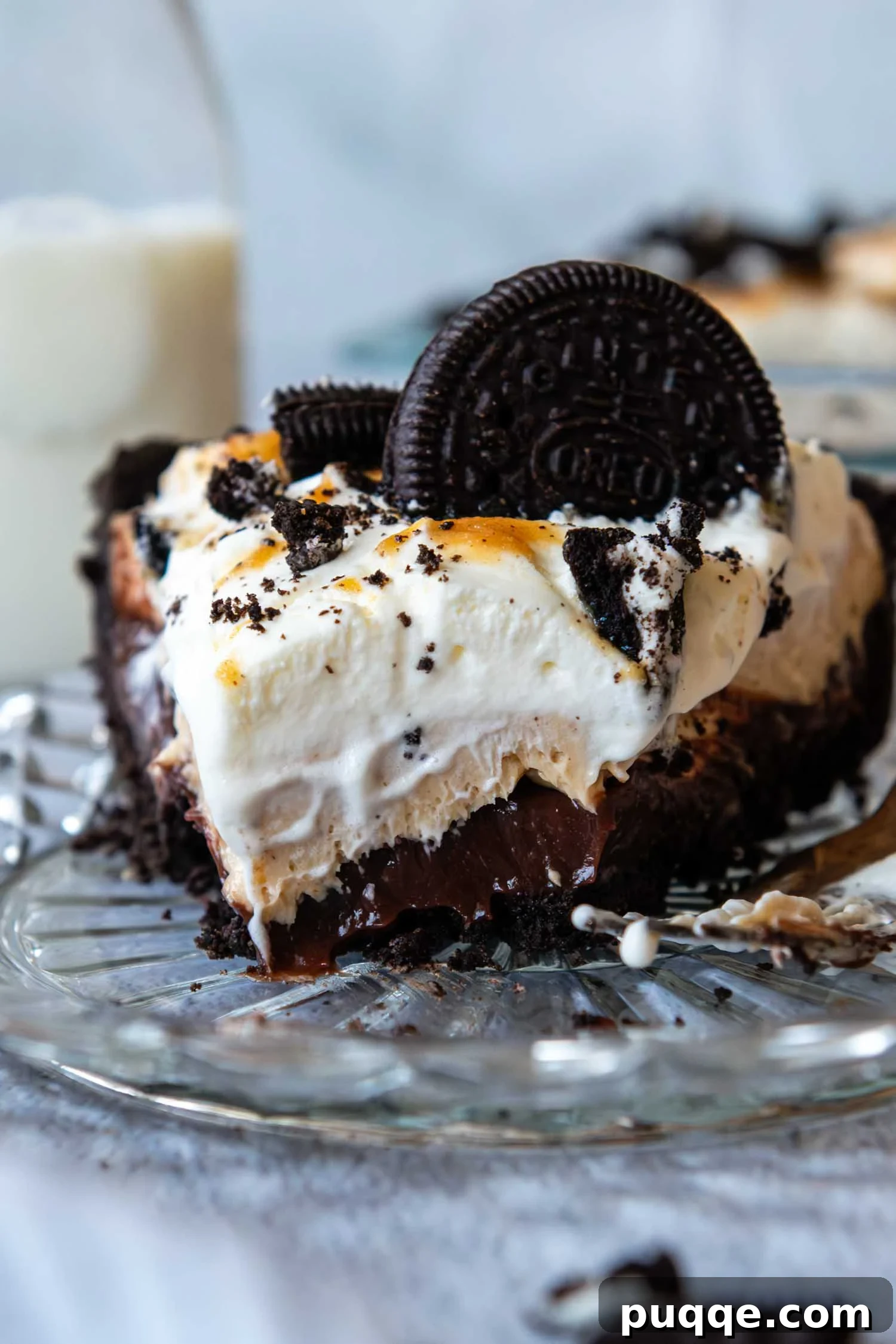 A delectable slice of peanut butter Oreo pie on a clear glass pie plate, topped with a mini Oreo cookie, showcasing its distinct layers of chocolate crust, rich pudding, creamy peanut butter filling, and fluffy whipped cream.