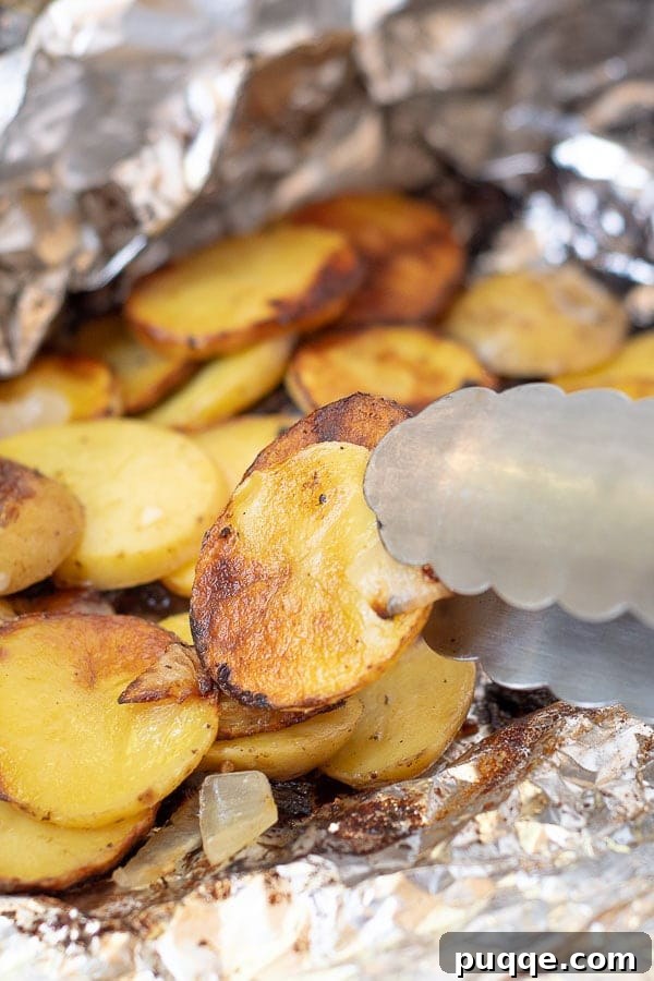 A pair of tongs lifting a perfectly grilled potato slice from an open foil packet, revealing its tender, cheesy goodness.