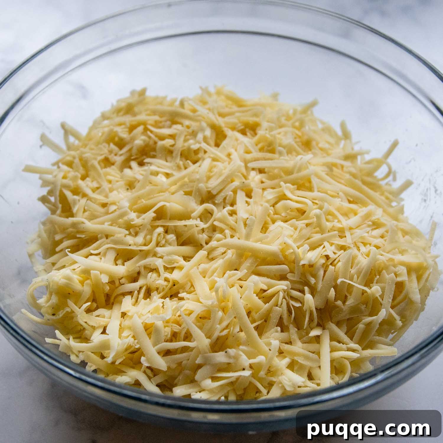Effortless Creamy Gluten-Free Cheese Fondue 3 A clear glass bowl brimming with freshly grated, golden Swiss cheese, seasoned and ready to be transformed into creamy fondue. The individual strands of cheese are visible, mixed with spices, awaiting their gentle heat bath.