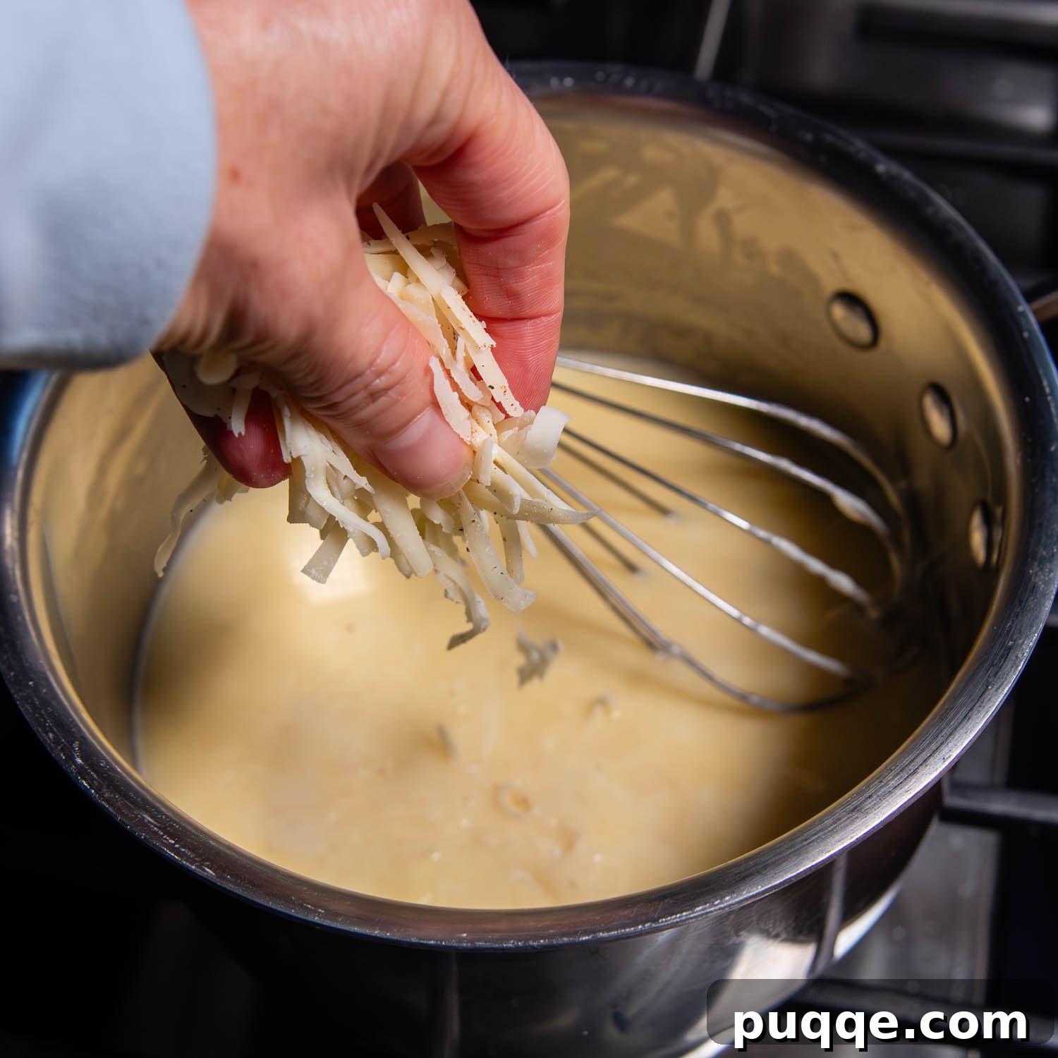Effortless Creamy Gluten-Free Cheese Fondue 5 A hand gently adds a handful of freshly grated Swiss cheese into a saucepan where wine is simmering, with a whisk ready to incorporate the cheese into the creamy fondue base.