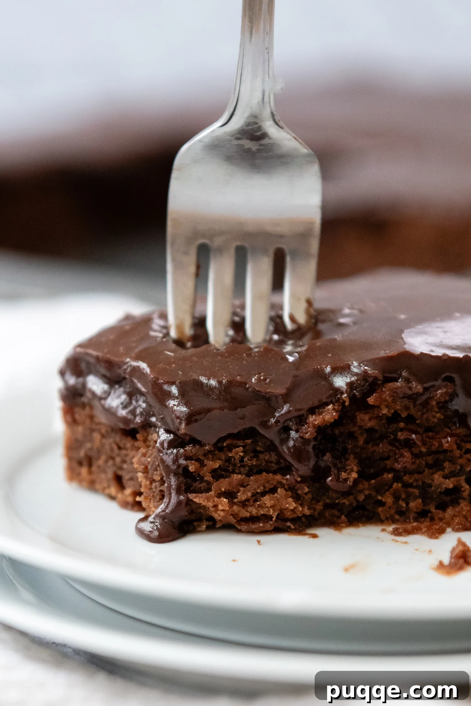 A delectable slice of Gluten-Free Texas Sheet Cake resting on a pristine white plate, accompanied by a fork. The cake features a rich chocolate hue and is topped with a smooth, glossy chocolate glaze, showcasing its tender crumb.