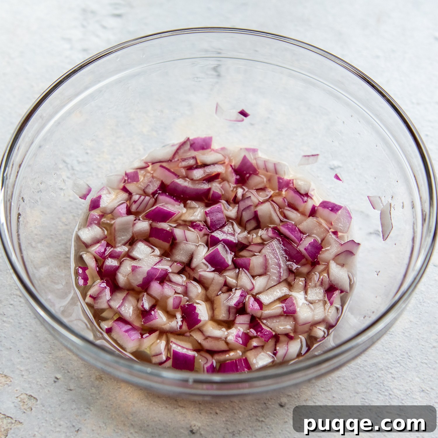 A glass bowl containing finely chopped red onions soaking in a pickling mixture, ready to infuse flavor into the salad.