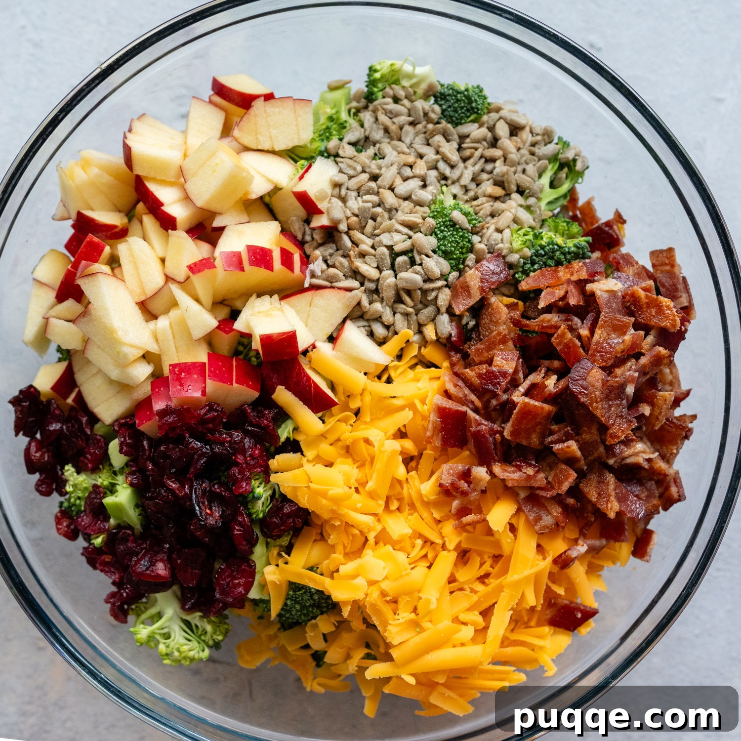 A large glass bowl filled with the colorful ingredients of the salad, including broccoli, cranberries, sunflower seeds, bacon, cheddar, and apple, ready for dressing.