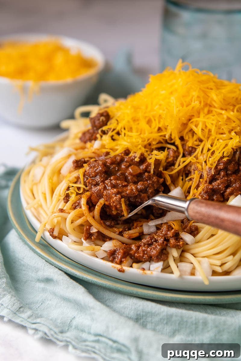 Ultimate Cincinnati Chili 6 A close-up of Cincinnati Chili served over spaghetti, generously topped with shredded cheese and a fork ready to dig in.