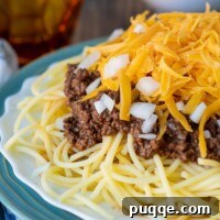 Ultimate Cincinnati Chili 8 A close-up of Cincinnati chili meat sauce on pasta with cheese, and a cup in the background.