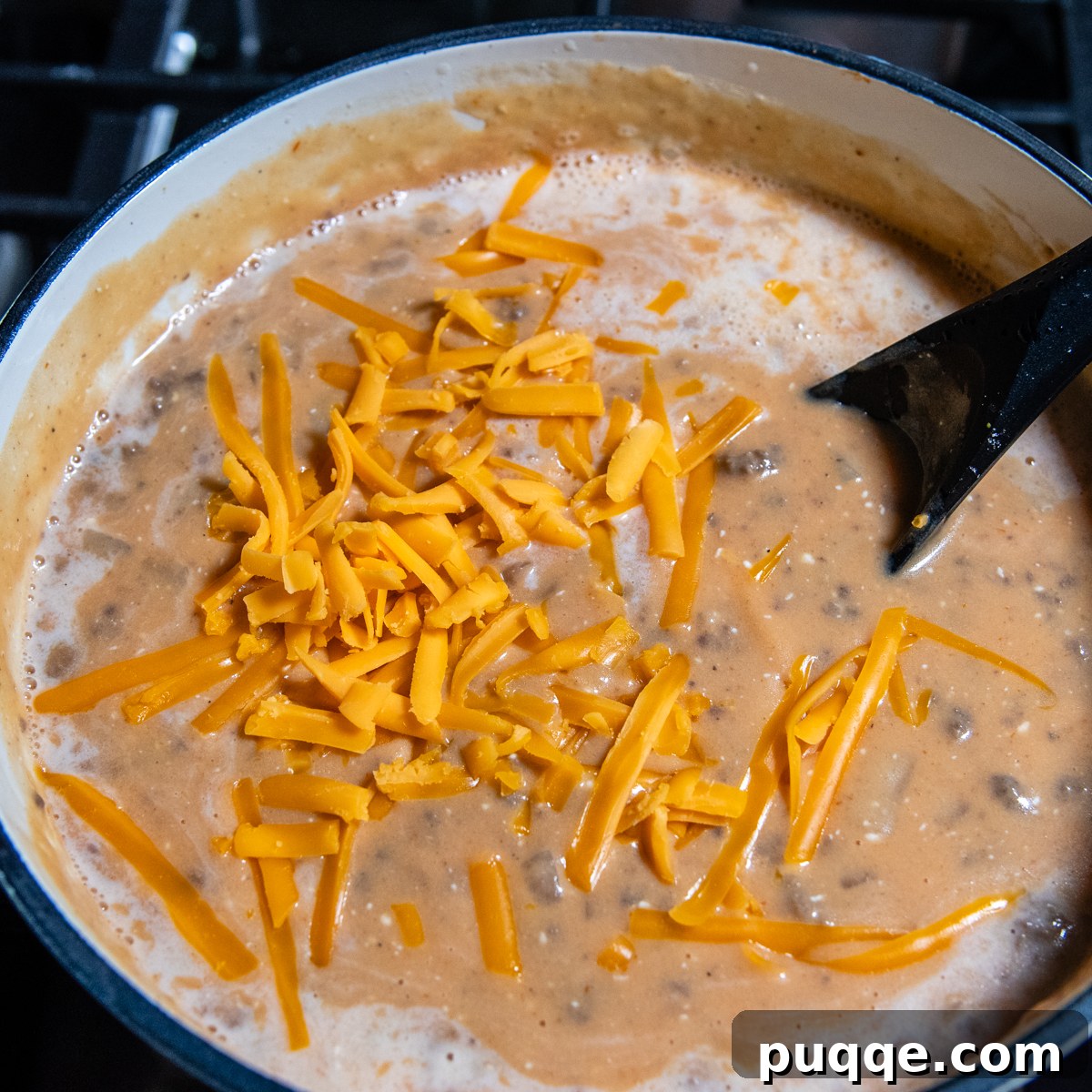 A black spoon stirring freshly grated cheddar cheese into the creamy cheeseburger soup.