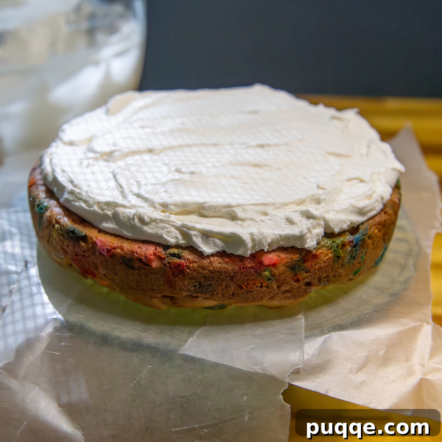 Fluffy Gluten-Free Funfetti Birthday Delight 7 A two-layer gluten-free funfetti cake on a cake stand with parchment paper underneath, being frosted with a spatula.