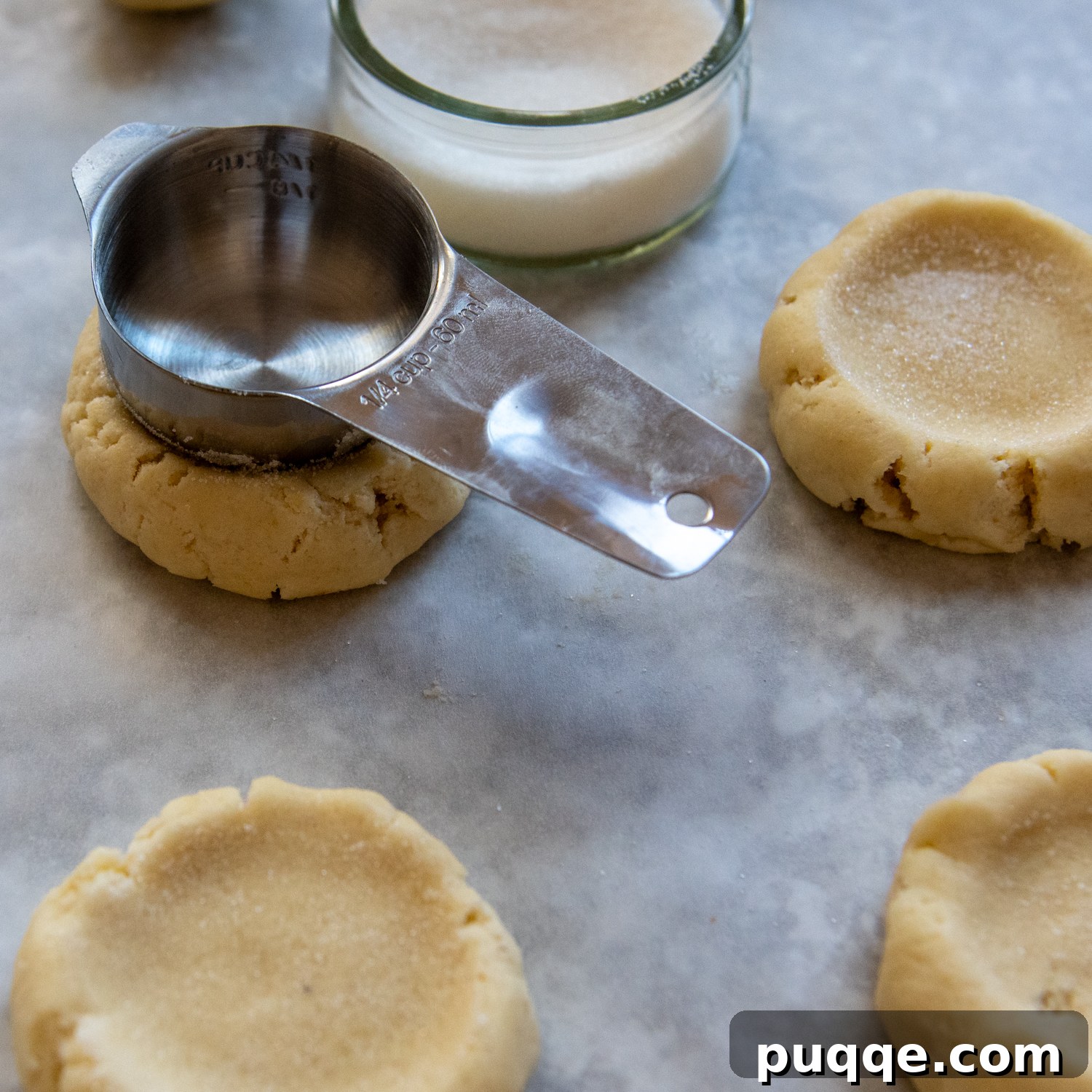 A measuring cup being carefully pressed into the center of a cookie dough ball to create a small indentation before baking.