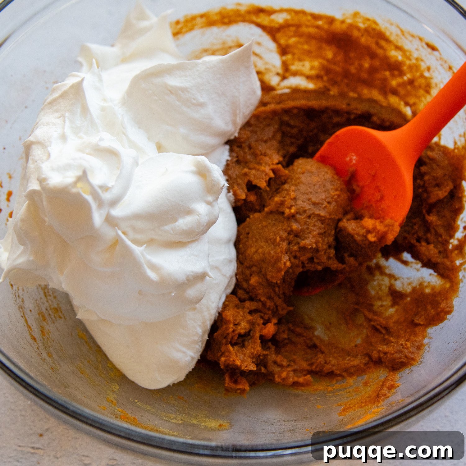 Frosting ingredients being gently combined with a red spatula in a clear glass mixing bowl, showcasing the creamy texture.