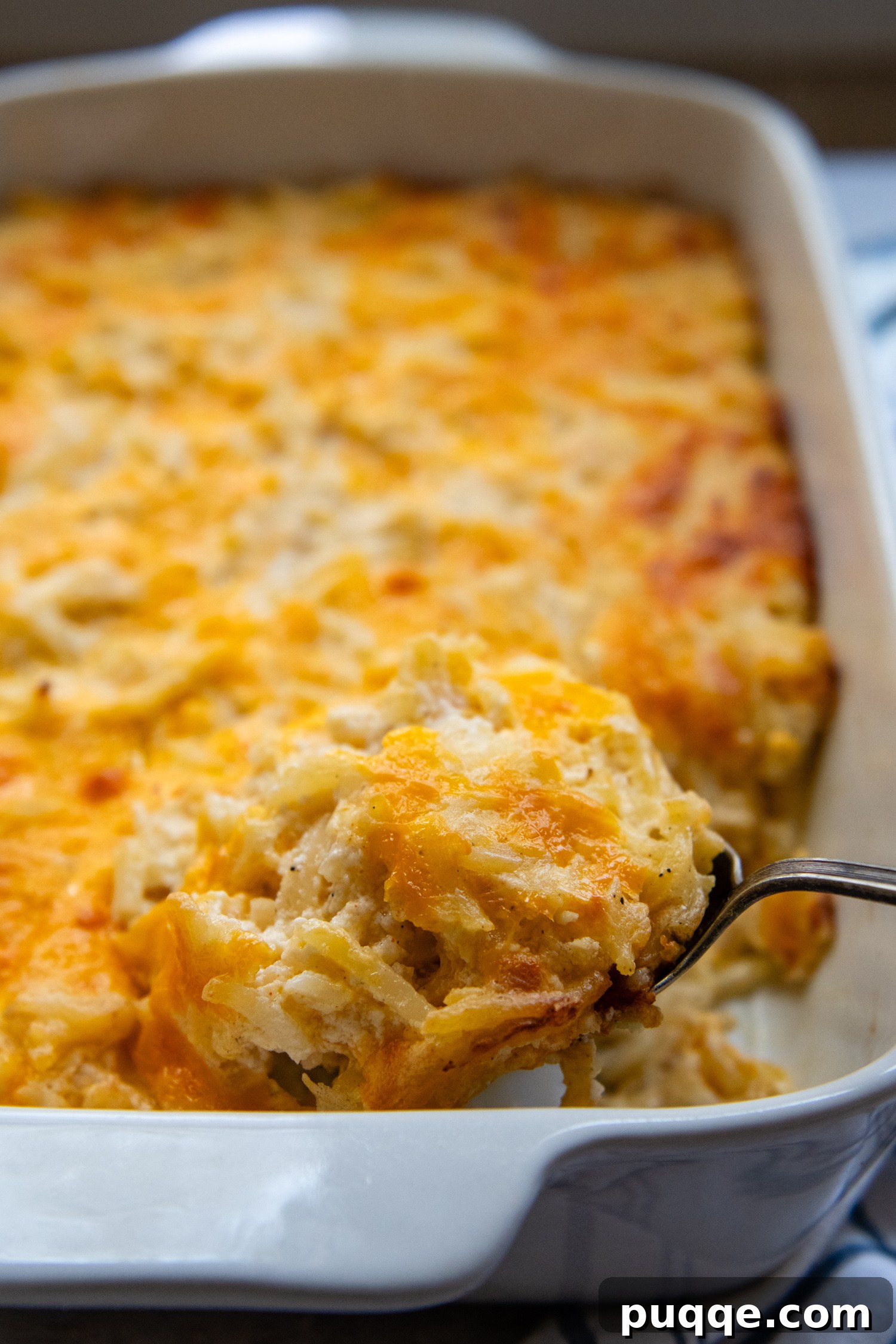 Ultimate Cheesy Gluten-Free Hash Brown Bake from Scratch 7 A scoop of rich, cheesy gluten-free hash brown casserole being lifted from a white baking dish, showcasing its creamy texture and golden brown top.