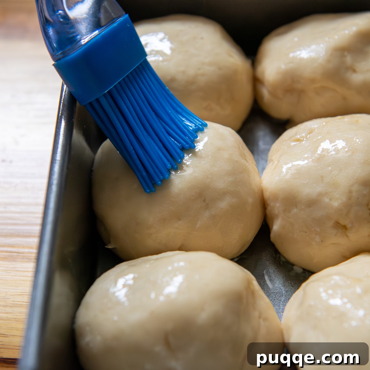 Gluten-Free Hawaiian Rolls: Squishy Soft in Just One Hour 7 A blue silicone brush applying melted butter to the tops of risen gluten-free Hawaiian rolls before baking.
