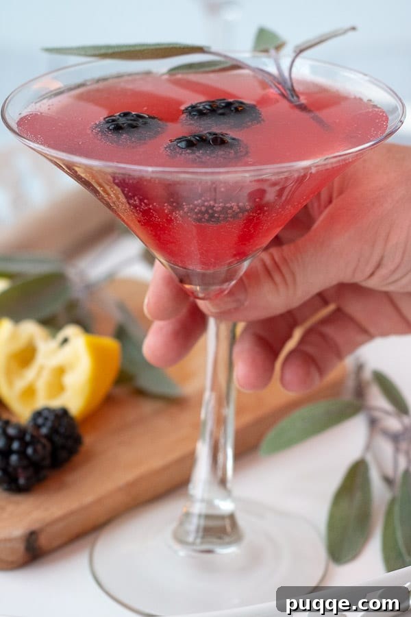 The Ultimate Blackberry Martini 2 A hand gently reaching for a martini glass filled with a sparkling blackberry martini, garnished with a fresh blackberry.