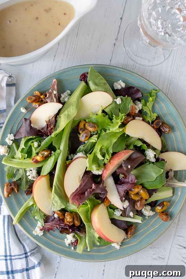 Overhead shot of a vibrant Apple Pecan Salad on a blue plate, with a glass of wine and a container of homemade Maple Dijon Vinaigrette nearby, evoking a warm autumn atmosphere.