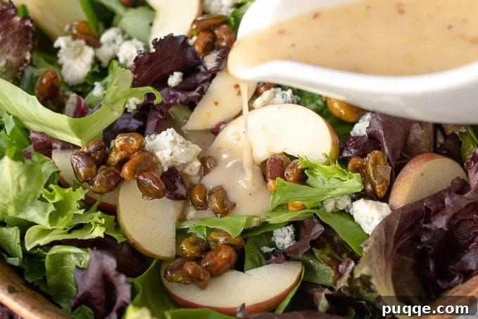 An inviting close-up shot of homemade Maple Dijon Vinaigrette being drizzled from a white dressing container onto the vibrant Apple Pecan Salad, highlighting the fresh ingredients.