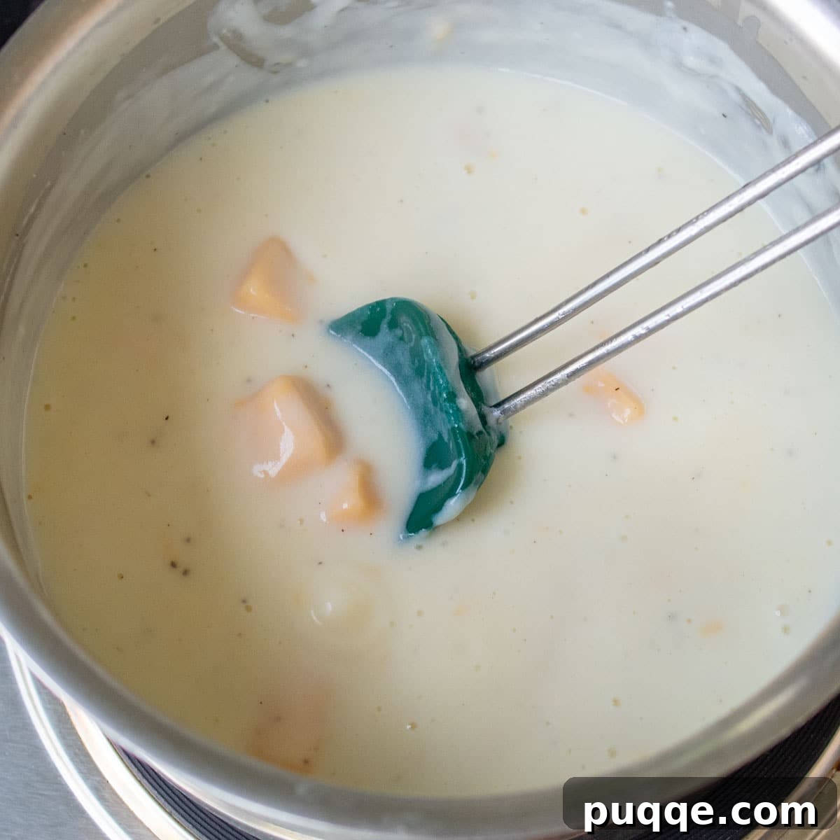 A creamy white sauce in a saucepan with chunks of Velveeta cheese just added, beginning to melt and blend into the sauce.