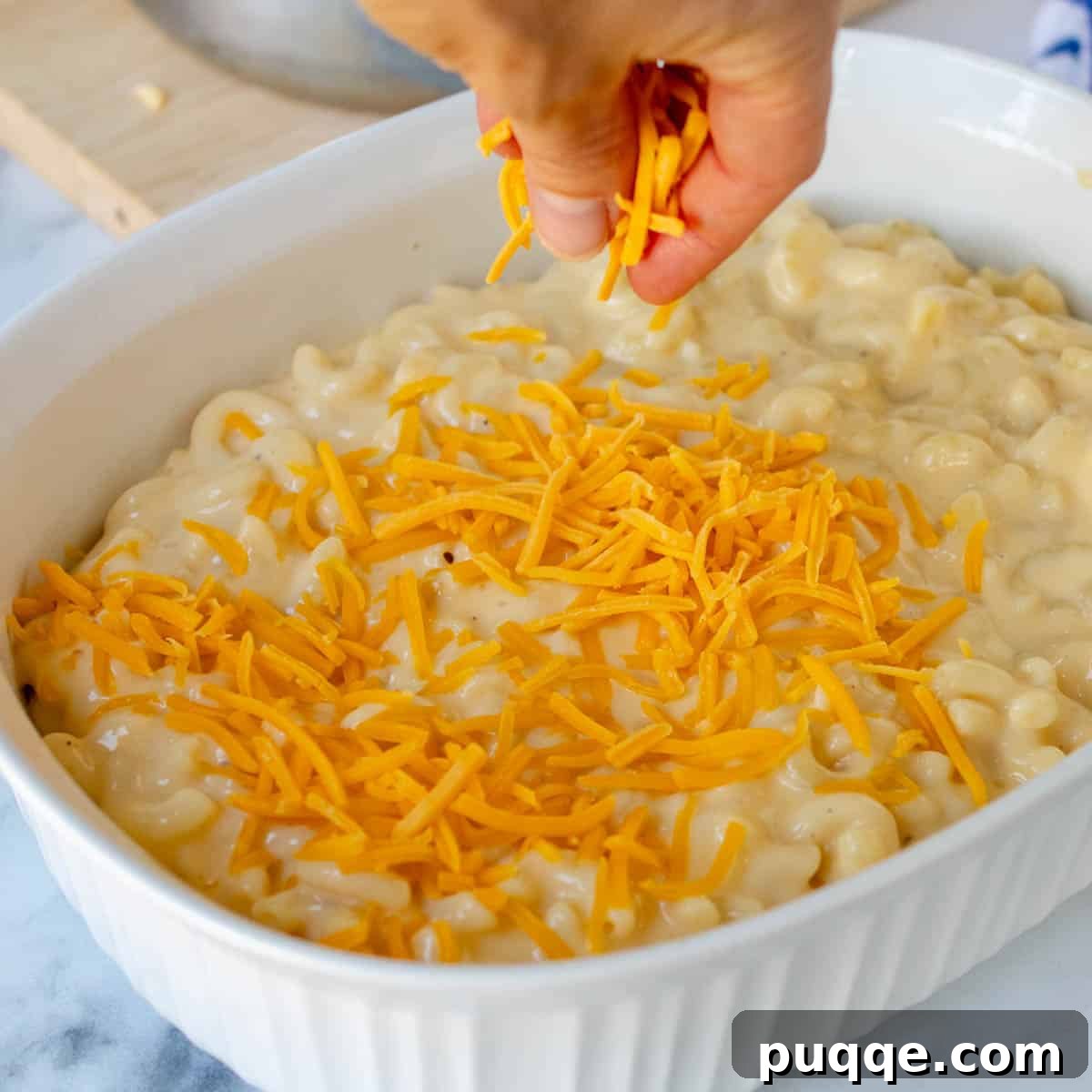 A casserole dish filled with mac and cheese, being topped with a generous layer of shredded cheddar cheese before going into the oven.