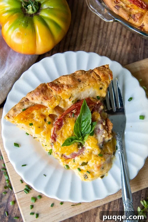 Easy Bisquick Tomato Pie 3 A perfectly portioned slice of tomato pie resting on a white scalloped plate, accompanied by a vibrant whole fresh tomato.