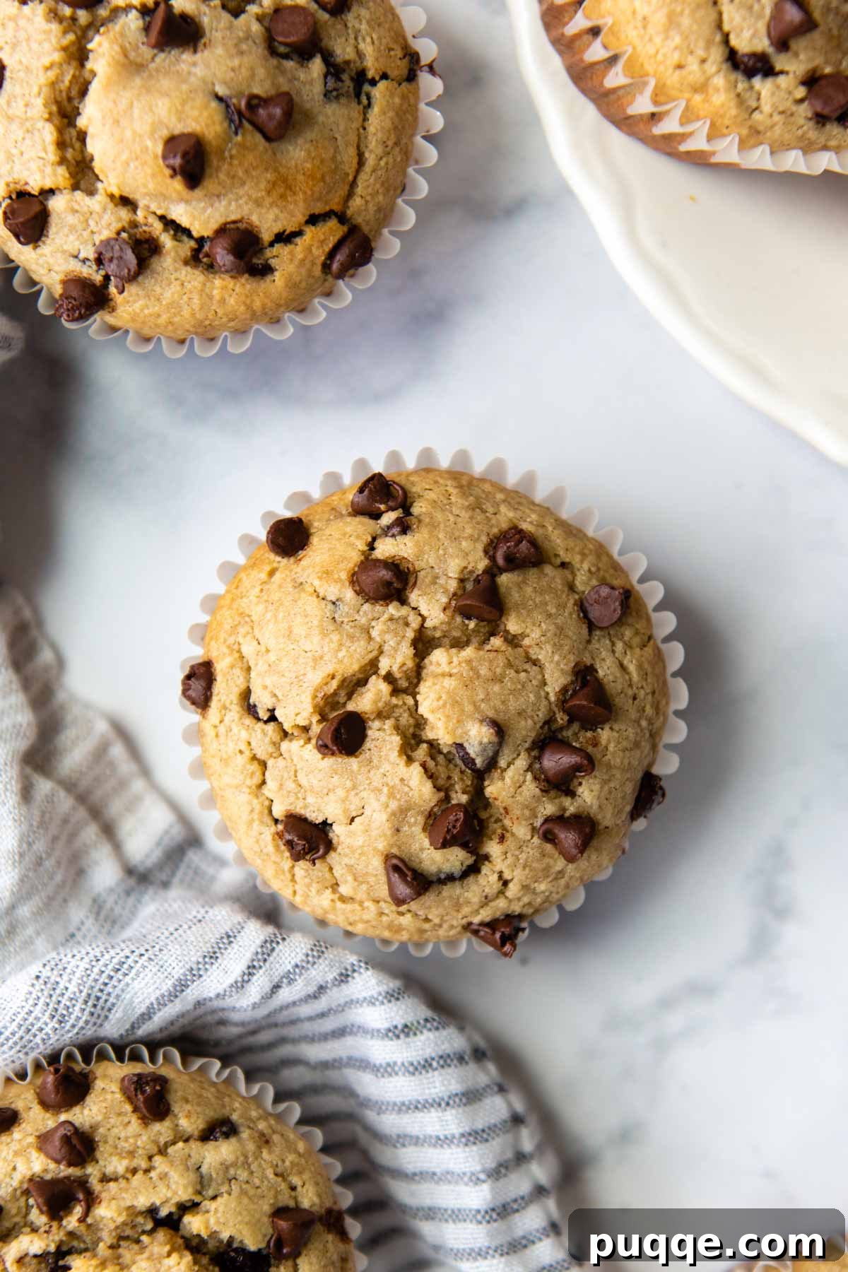 A top-down shot of perfectly baked almond flour chocolate chip muffins arranged on a pristine white marble surface, accompanied by a soft gray linen napkin.