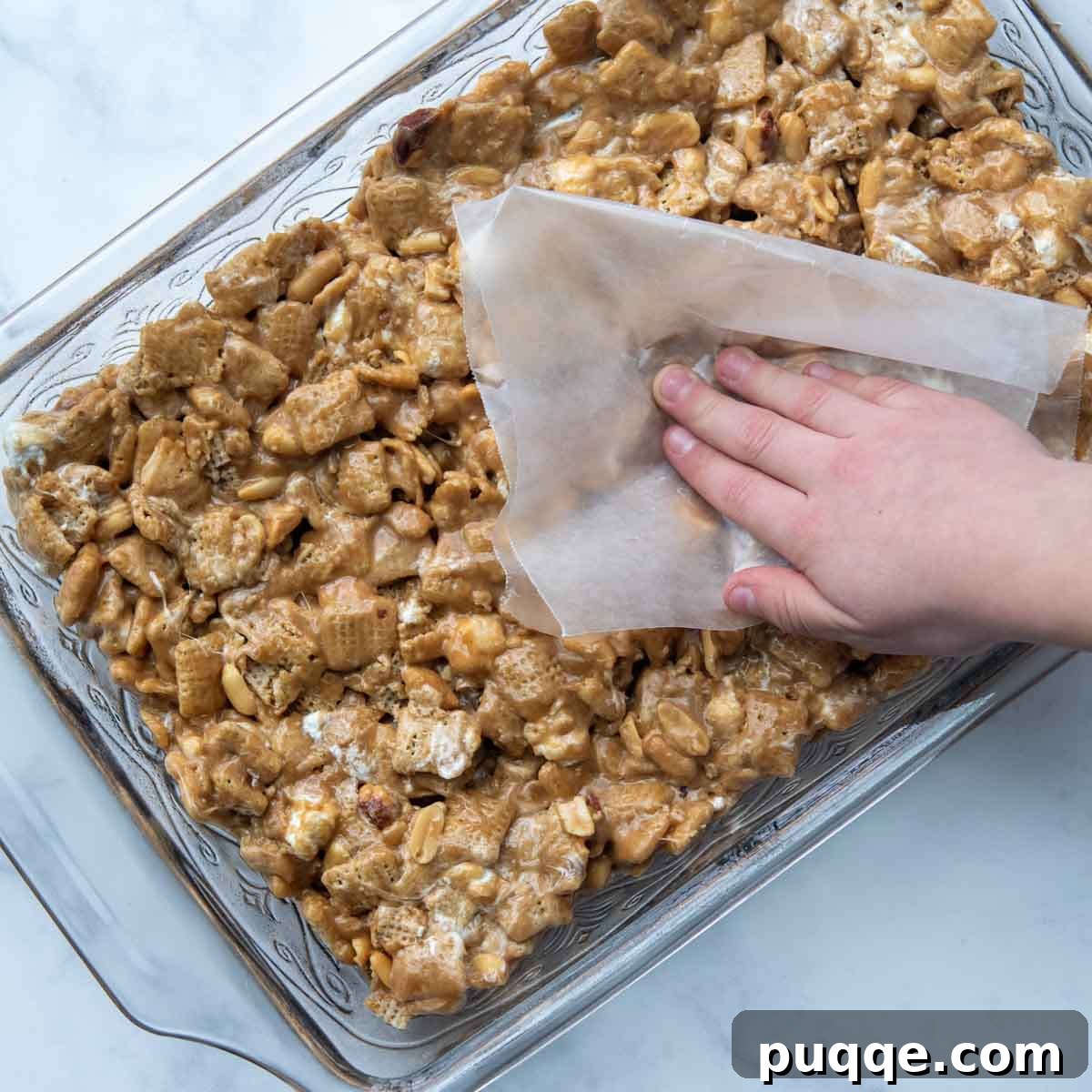 A piece of greased waxed paper being used to gently and evenly press down the Scotcheroos mixture in a baking dish, ensuring a smooth top layer without compacting the bars.