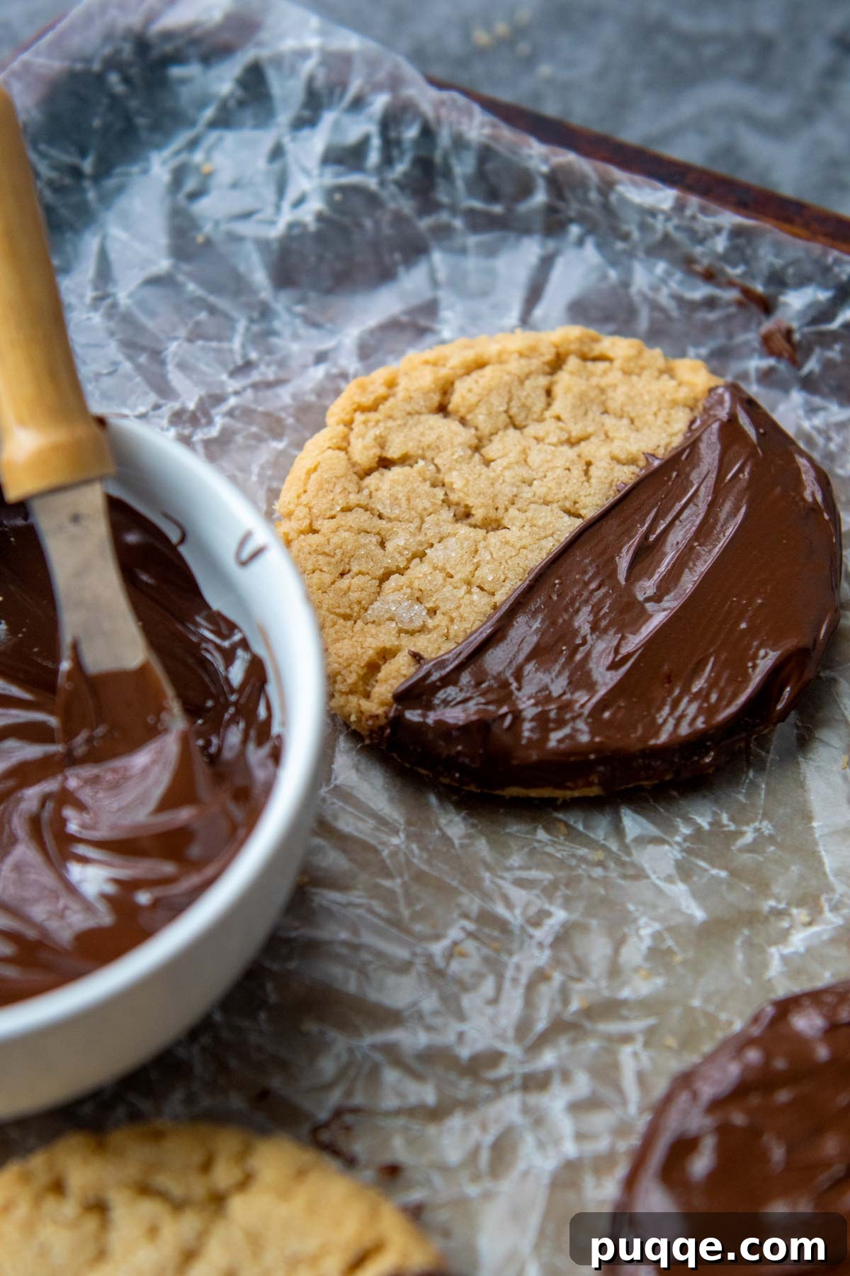 A freshly baked almond flour peanut butter cookie partially dipped in rich, melted chocolate, with a bowl of melted chocolate next to it.
