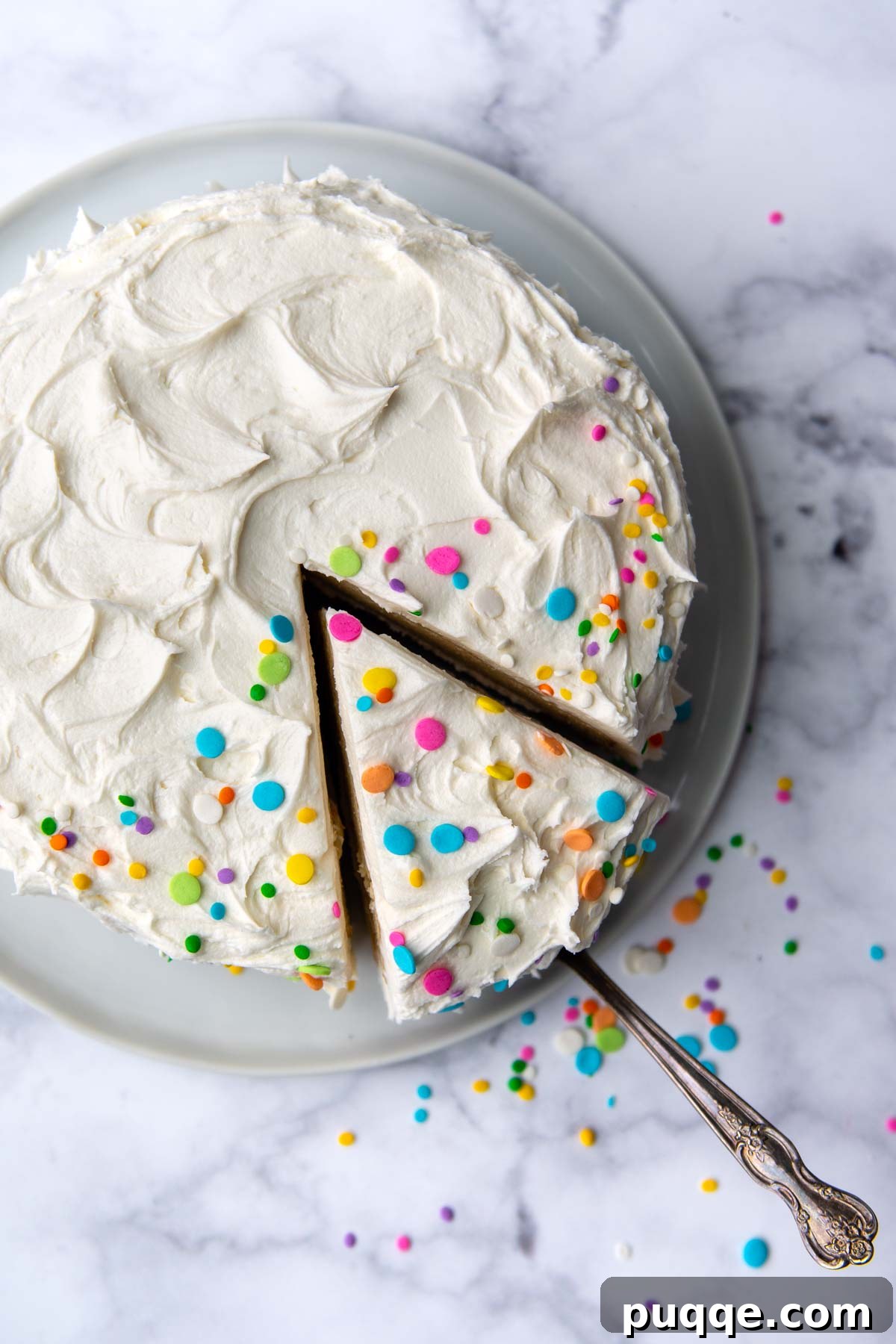Overhead shot of a hand taking a piece of frosted gluten-free white cake from a whole cake on a pedestal.