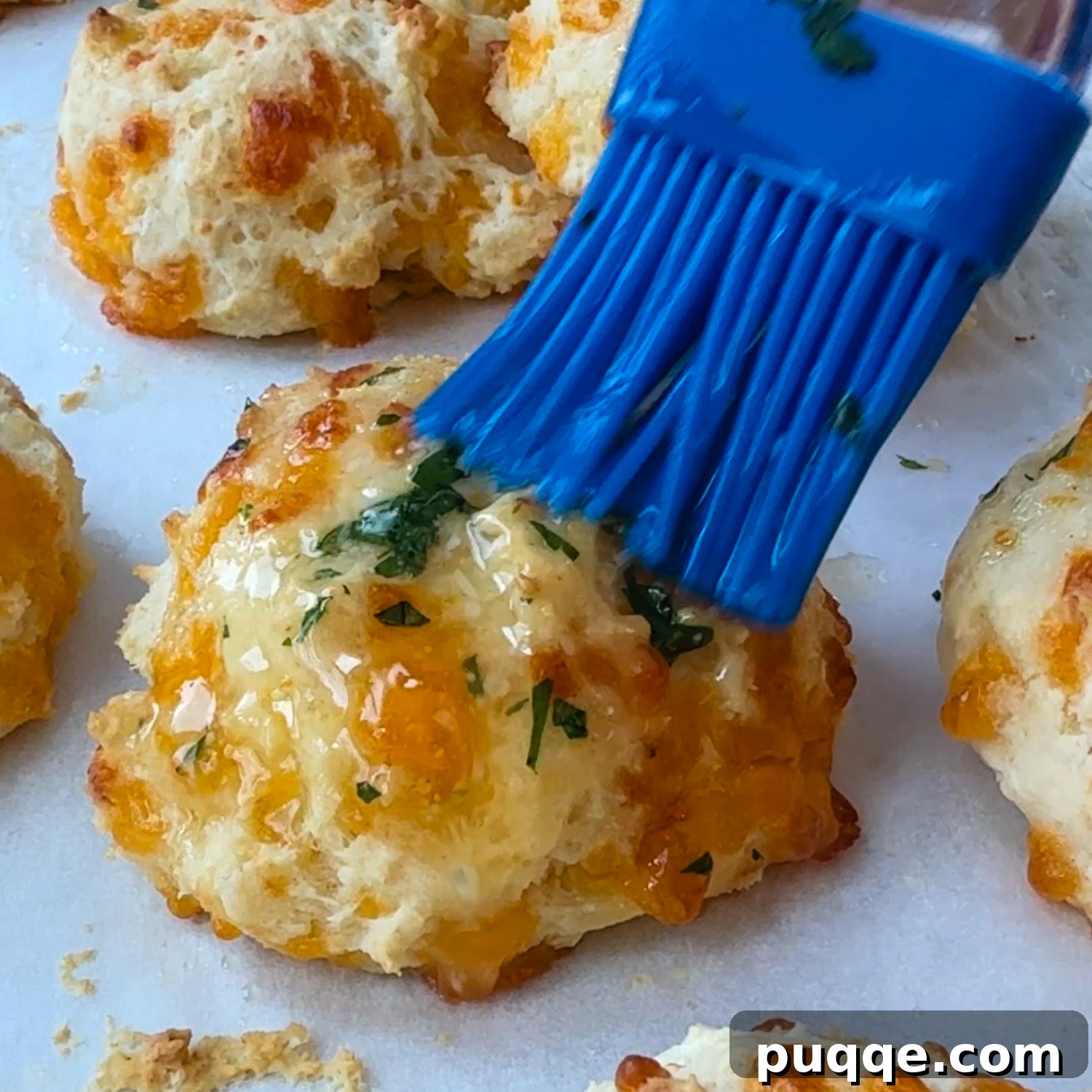Restaurant Style Buttery Gluten-Free Cheddar Bay Biscuits 6 A blue basting brush generously spreading melted garlic butter on a tray of freshly baked, golden-brown biscuits.
