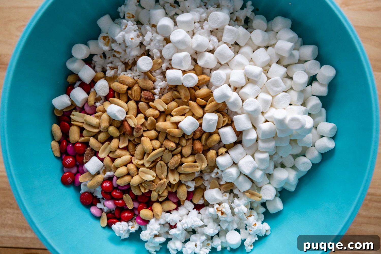 A large teal bowl brimming with a colorful medley of unadorned popcorn, salted peanuts, M&Ms, and fluffy mini marshmallows, ready for the white chocolate coating.