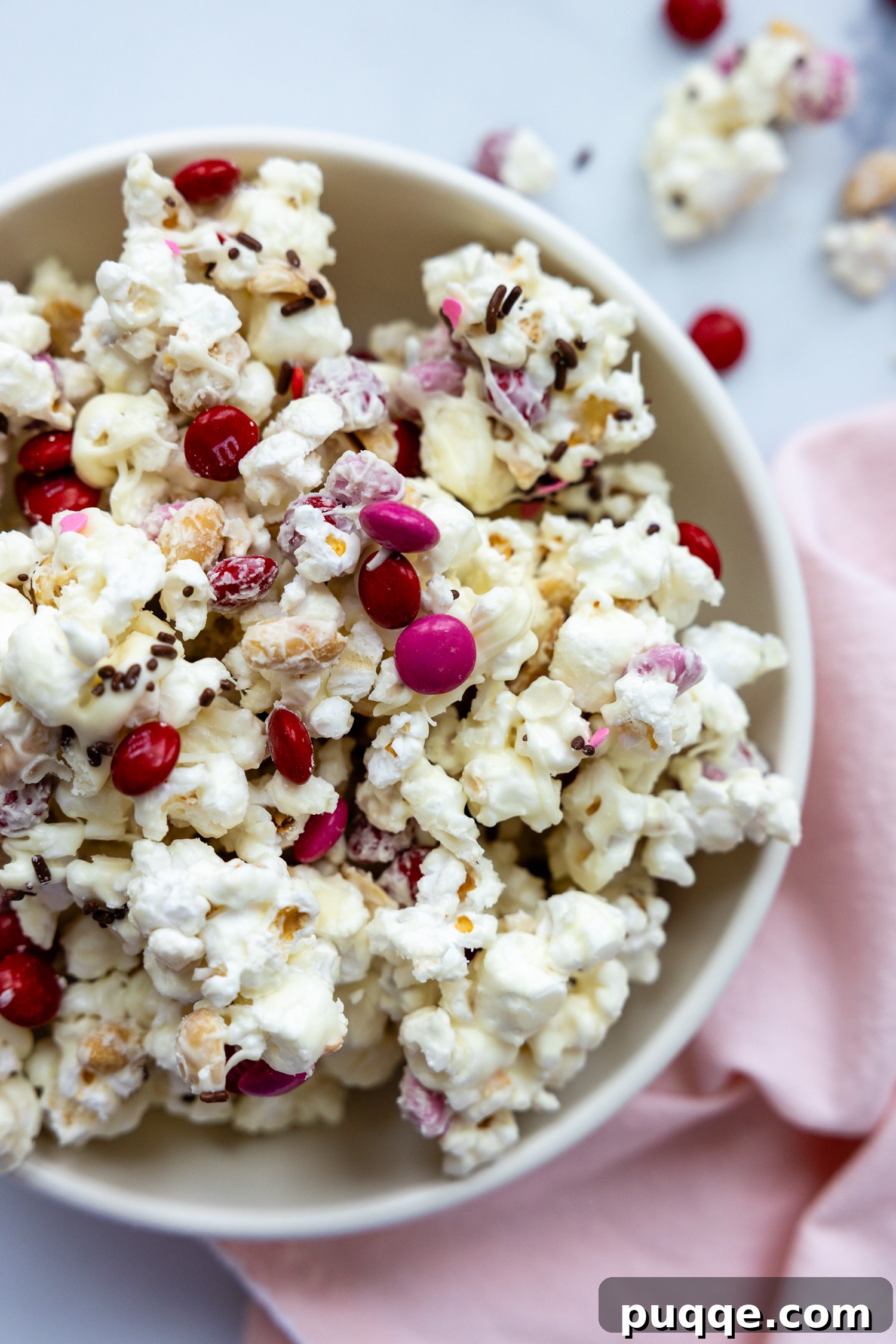 A inviting bowl of white chocolate popcorn mix, generously coated and adorned with M&Ms, resting beside a soft pink towel, showcasing its irresistible texture.
